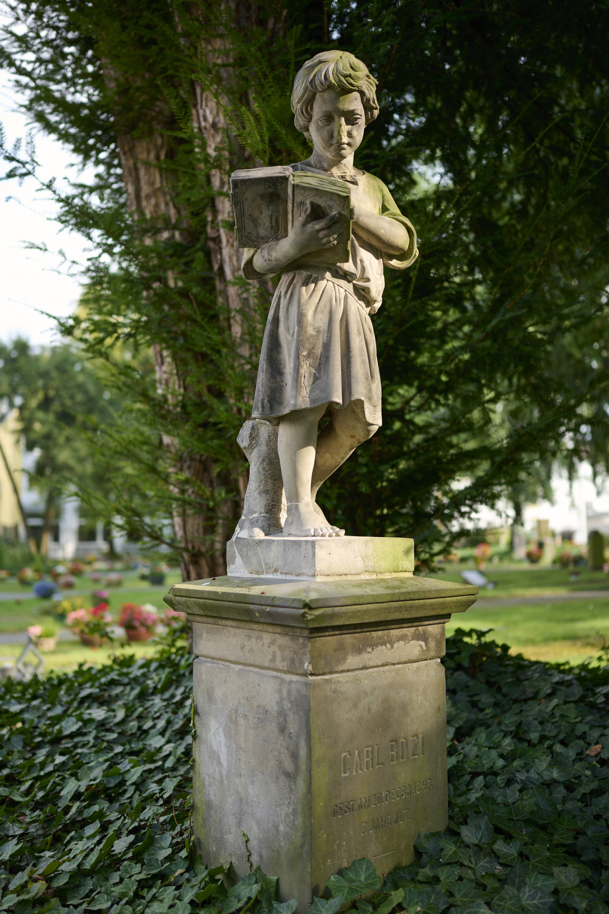 Bildtitel - Grabstätte von Carl Bozi: Eine Skulptur eines Jungen in einem gegürteten Kleid, der aus einem Buch liest.