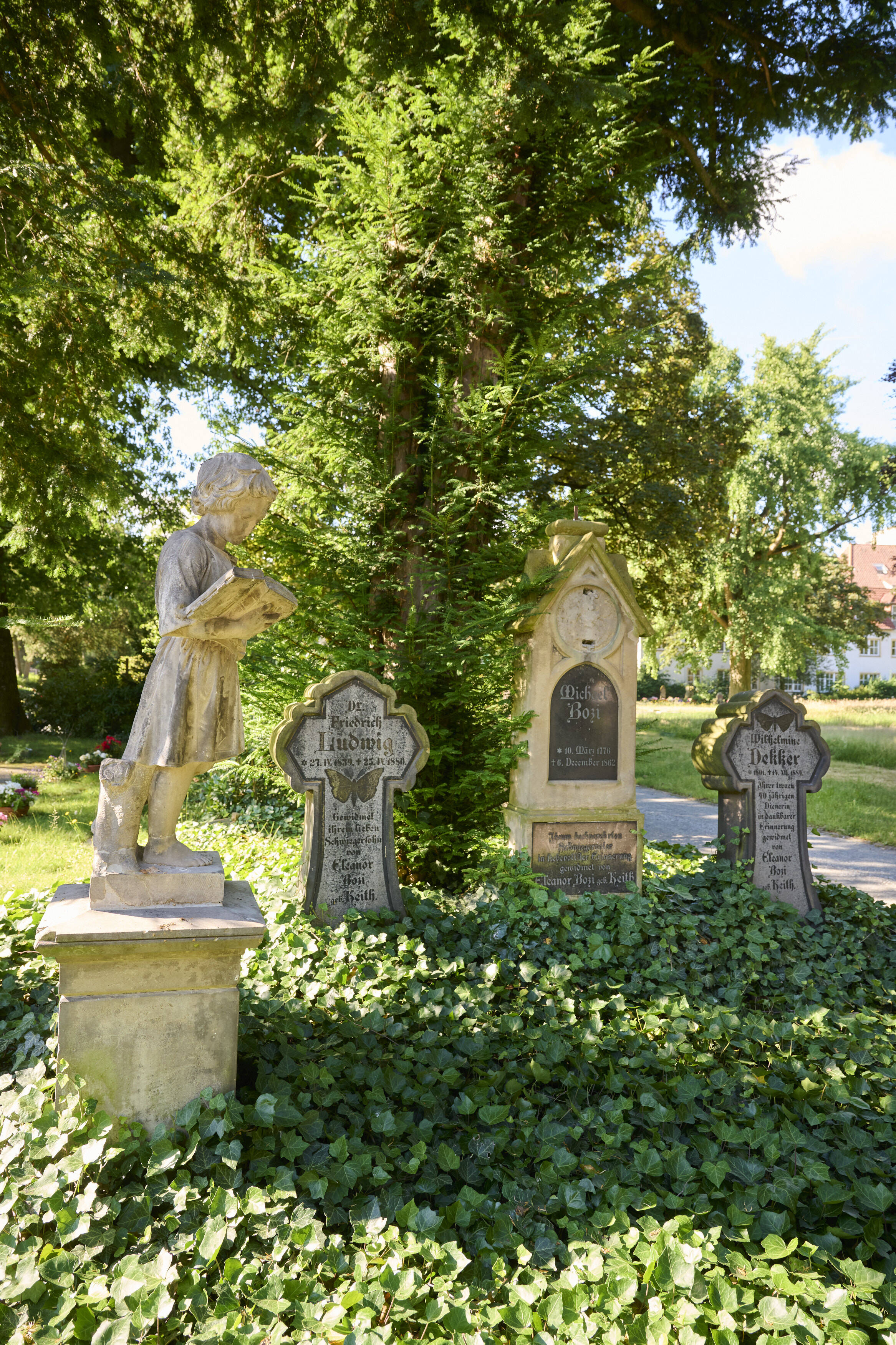 Bildtitel - Grabstätte von Michael Bozi: Zwei kreuzförmige Grabsteine mit Schmetterlings-Verzierung umrahmen einen größeren, nach oben spitz zulaufenden Grabstein. Links im Bild steht die Skulptur eines lesenden Jungen zu Ehren Carl Bozis.